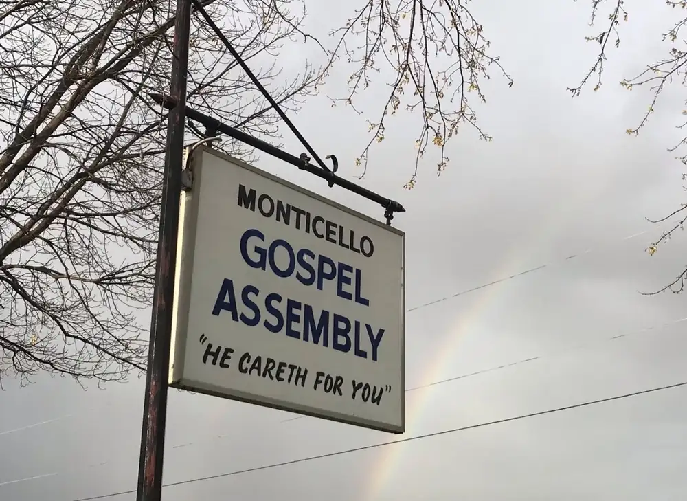 The church sign reading Monticello Gospel Assembly, He careth for you, with a rainbow behind it